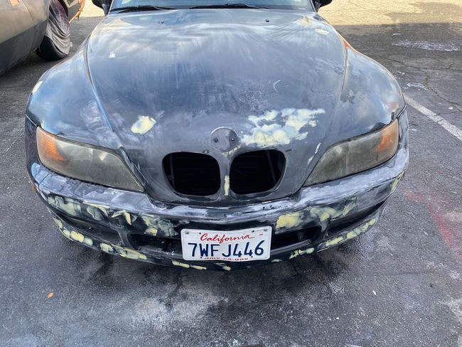 Damaged black BMW sedan with chipped hood and bumper, parked on frosty pavement.