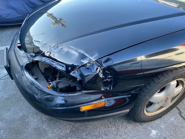 Black car with severe front-left damage, crumpled hood, and broken headlight after a crash