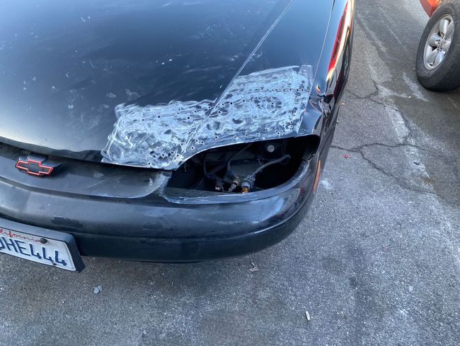 Damaged black car front corner with shattered windshield and broken headlight on pavement