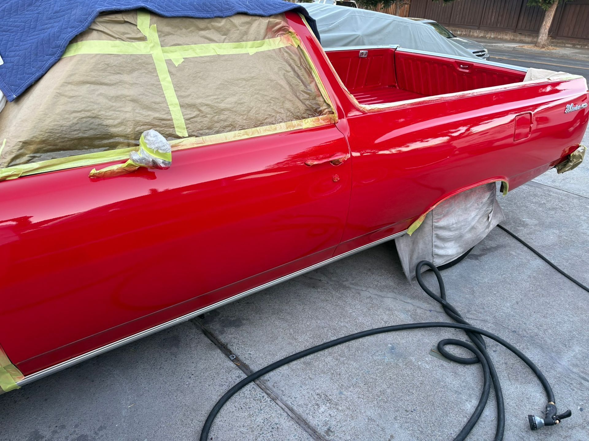 Red classic car under restoration, masked windows and taped trim, with a hose on the pavement.