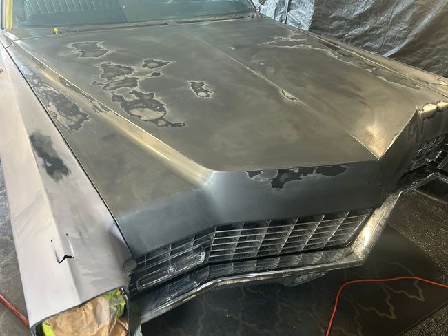Damaged gray car hood with dents and peeling paint, front grille visible in a workshop setting