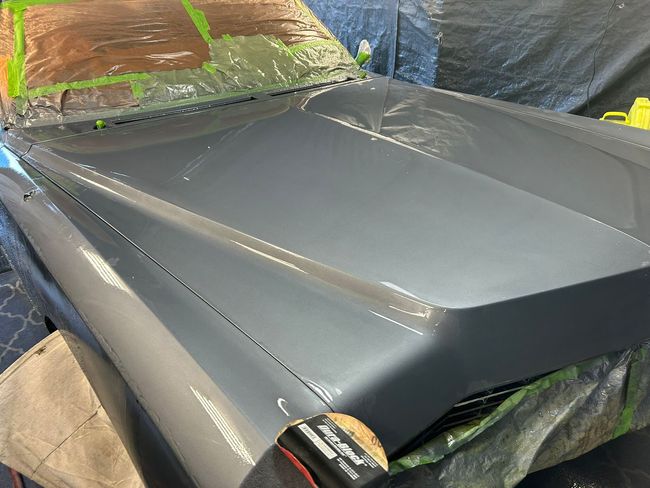 Gray car hood being painted in a workshop, masked with tape and plastic sheets