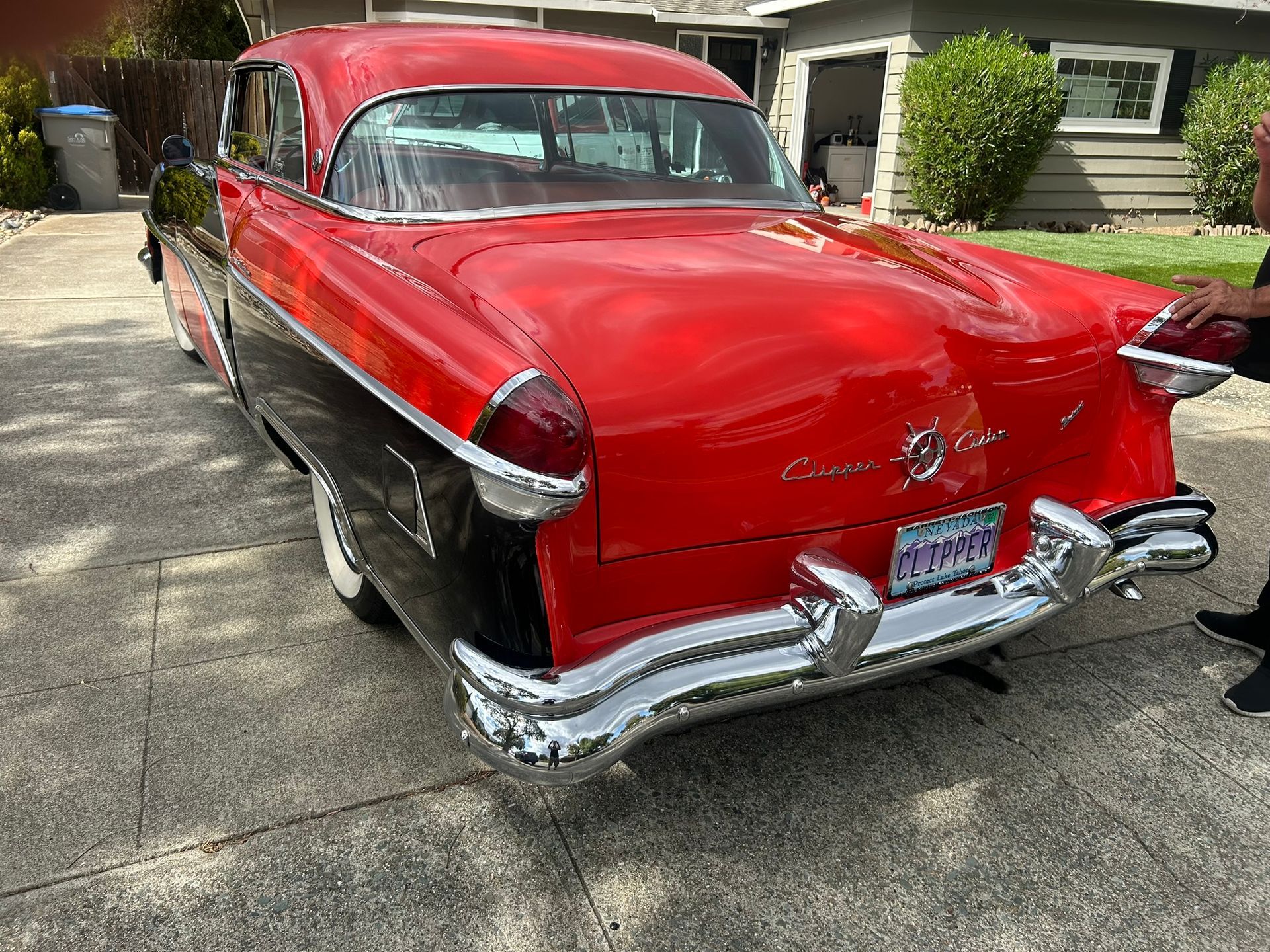 Red classic car parked in a driveway beside a house, rear view with chrome bumper.