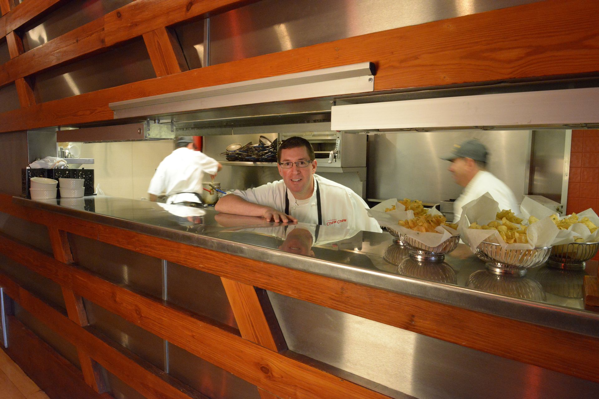 Chef leaning on counter in a restaurant kitchen, smiling at camera; other chefs working in background.