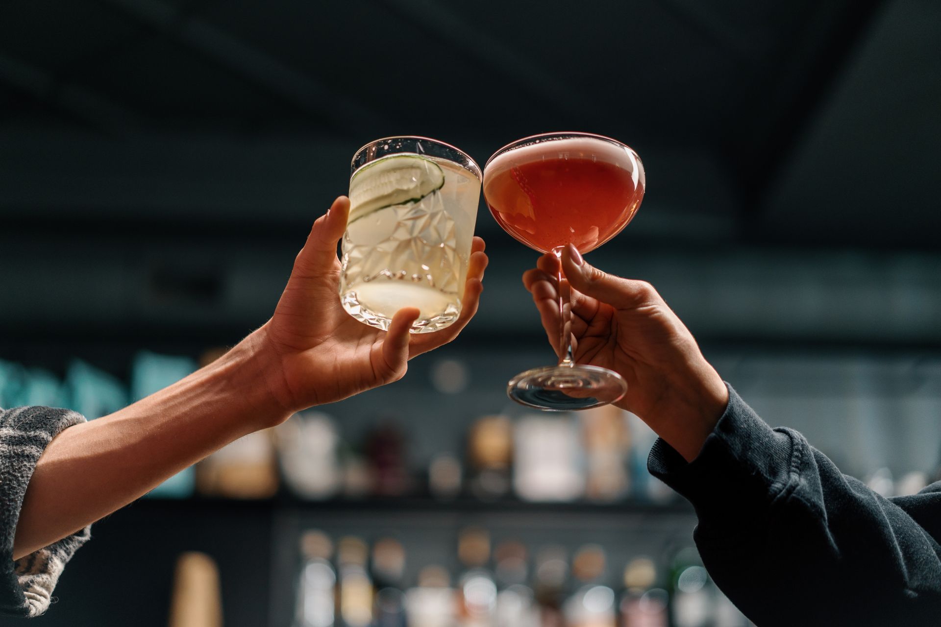 Two people toasting cocktails at a bar; one is clear with cucumber, the other is pink.