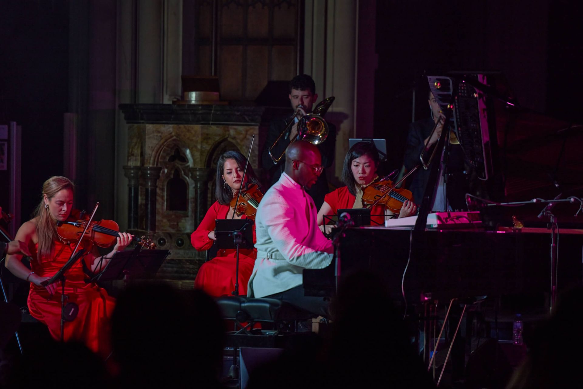 An orchestra performing onstage. A pianist in a white jacket surrounded by violinists in red dresses.
