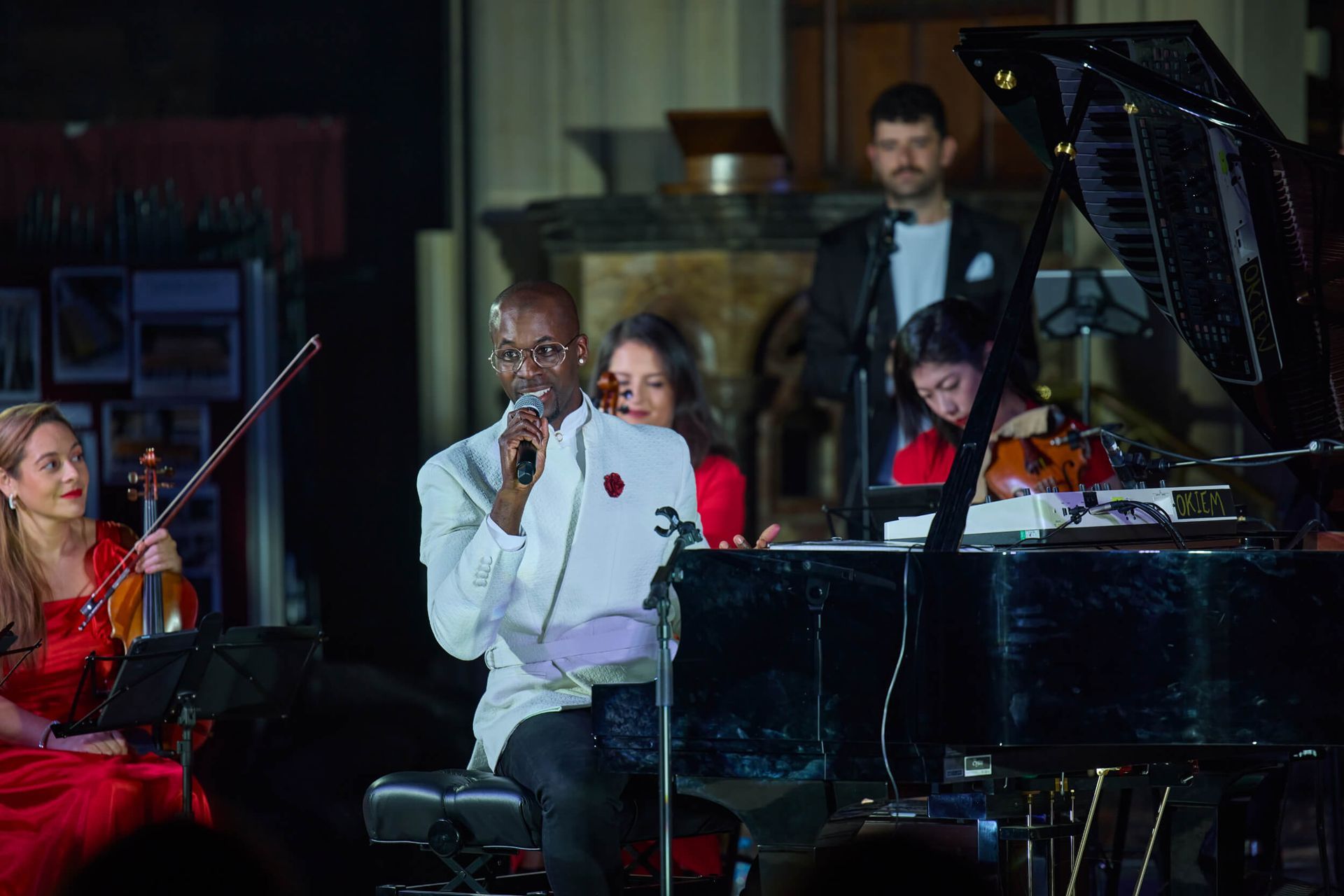 Musical ensemble performing: vocalist with microphone, piano, violins, and drummer in dimly lit room.