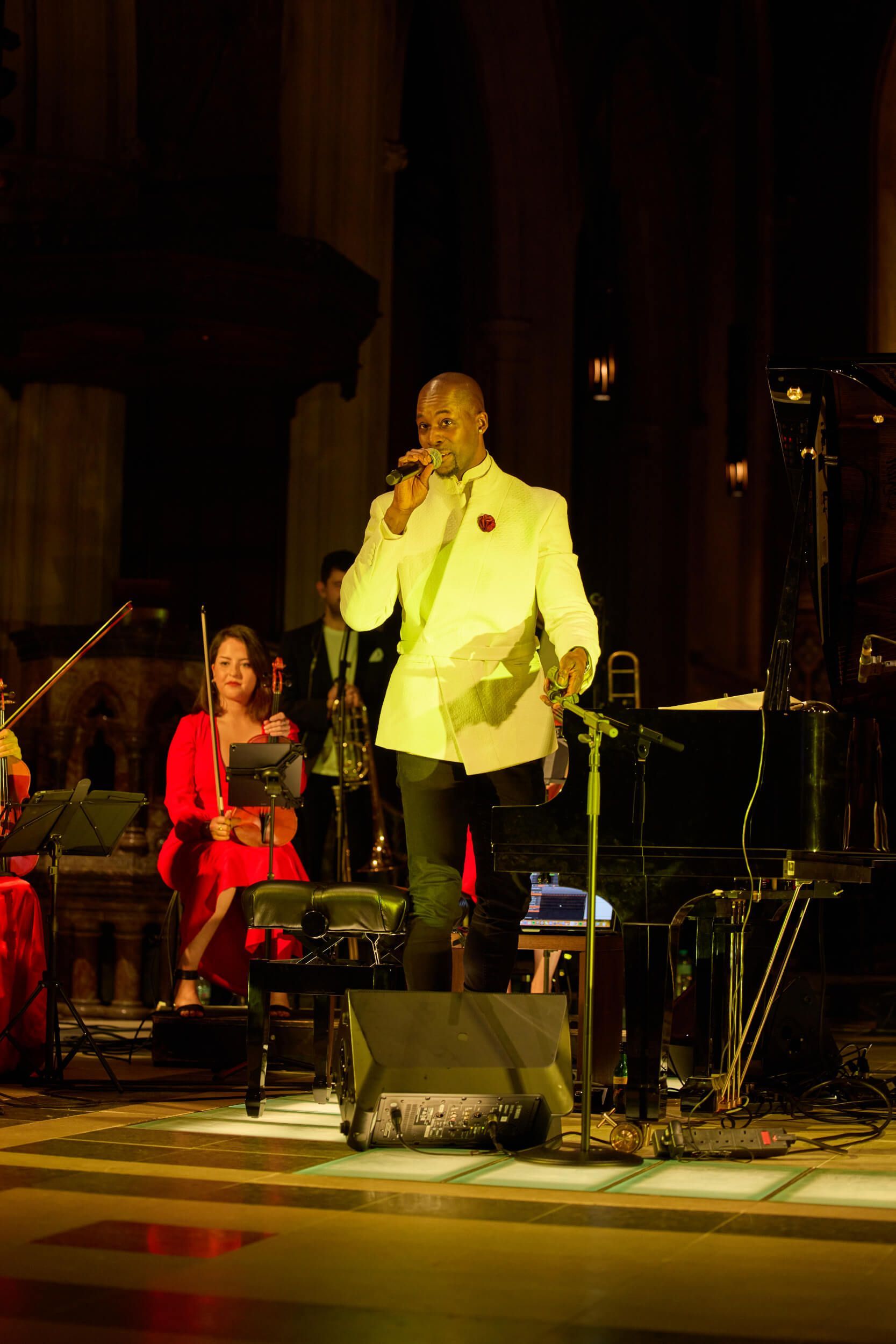 Man in white suit singing on stage, holding microphone. Band and piano in the background.
