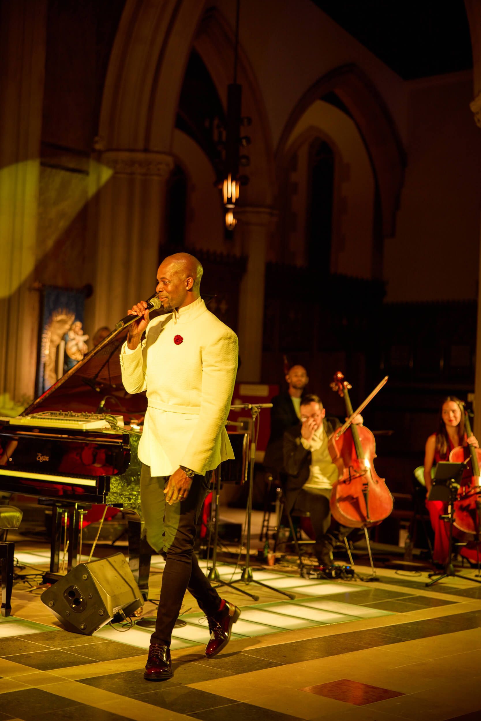 Singer in white jacket performs on stage with band, piano, and cello in a church.