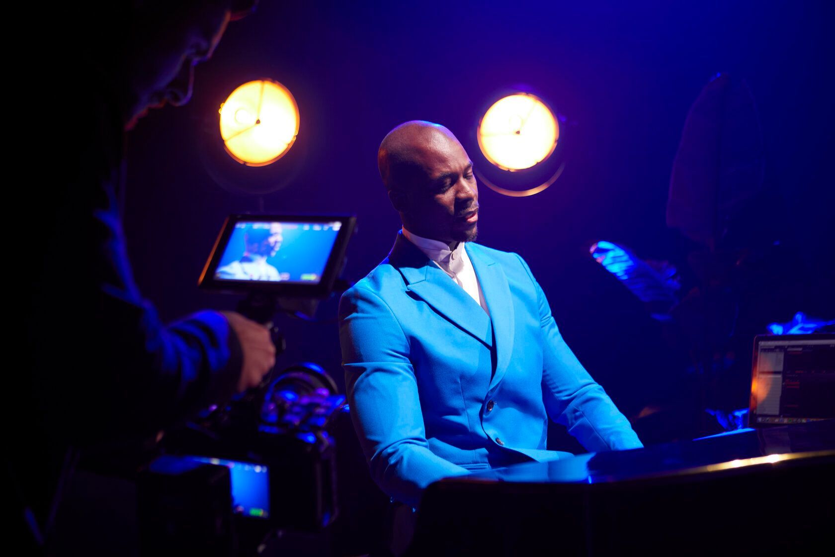 Man in blue suit, playing keys in a dark room, illuminated by stage lights.