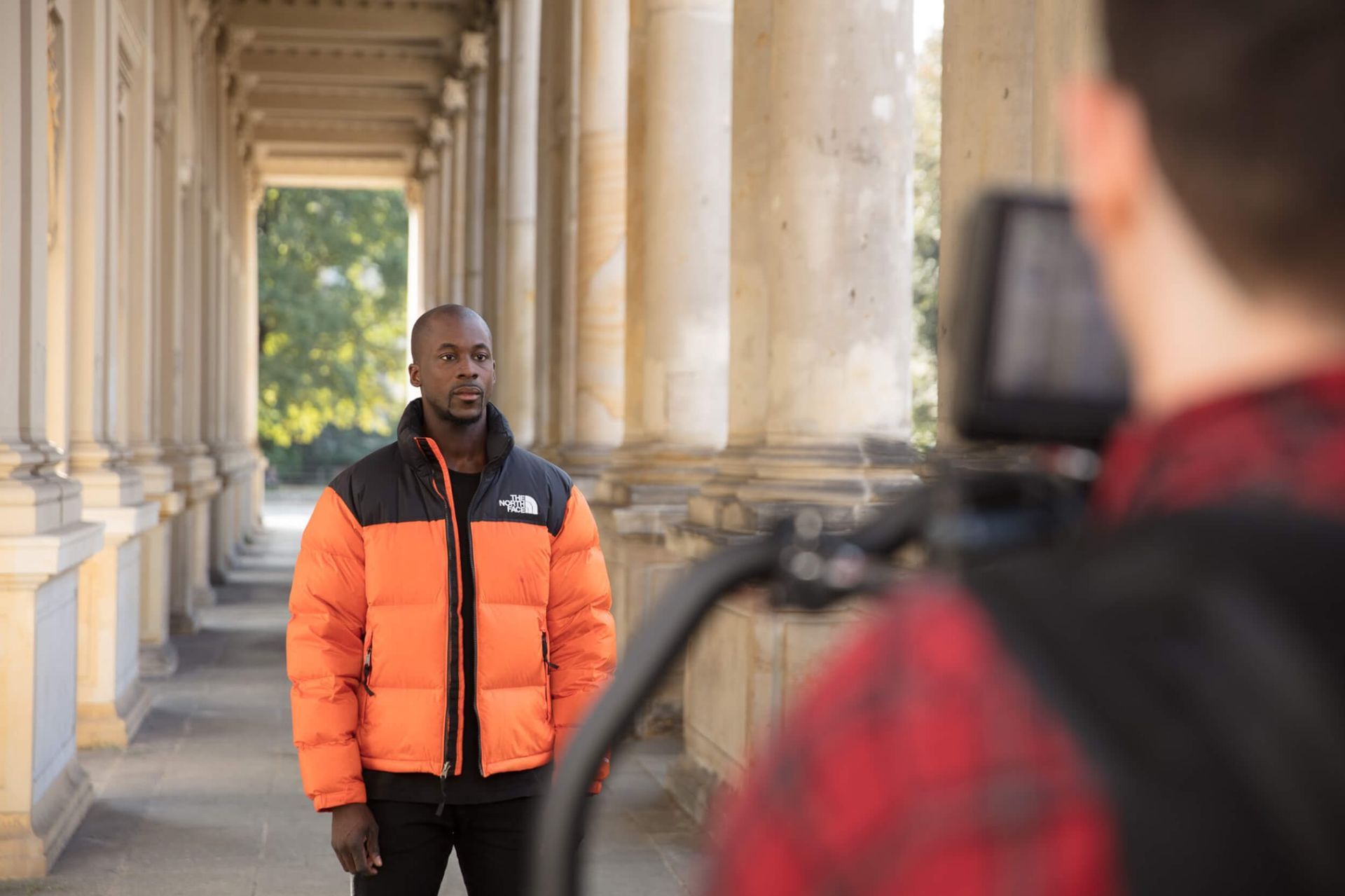 Man in orange jacket being filmed outdoors, standing in front of columns.