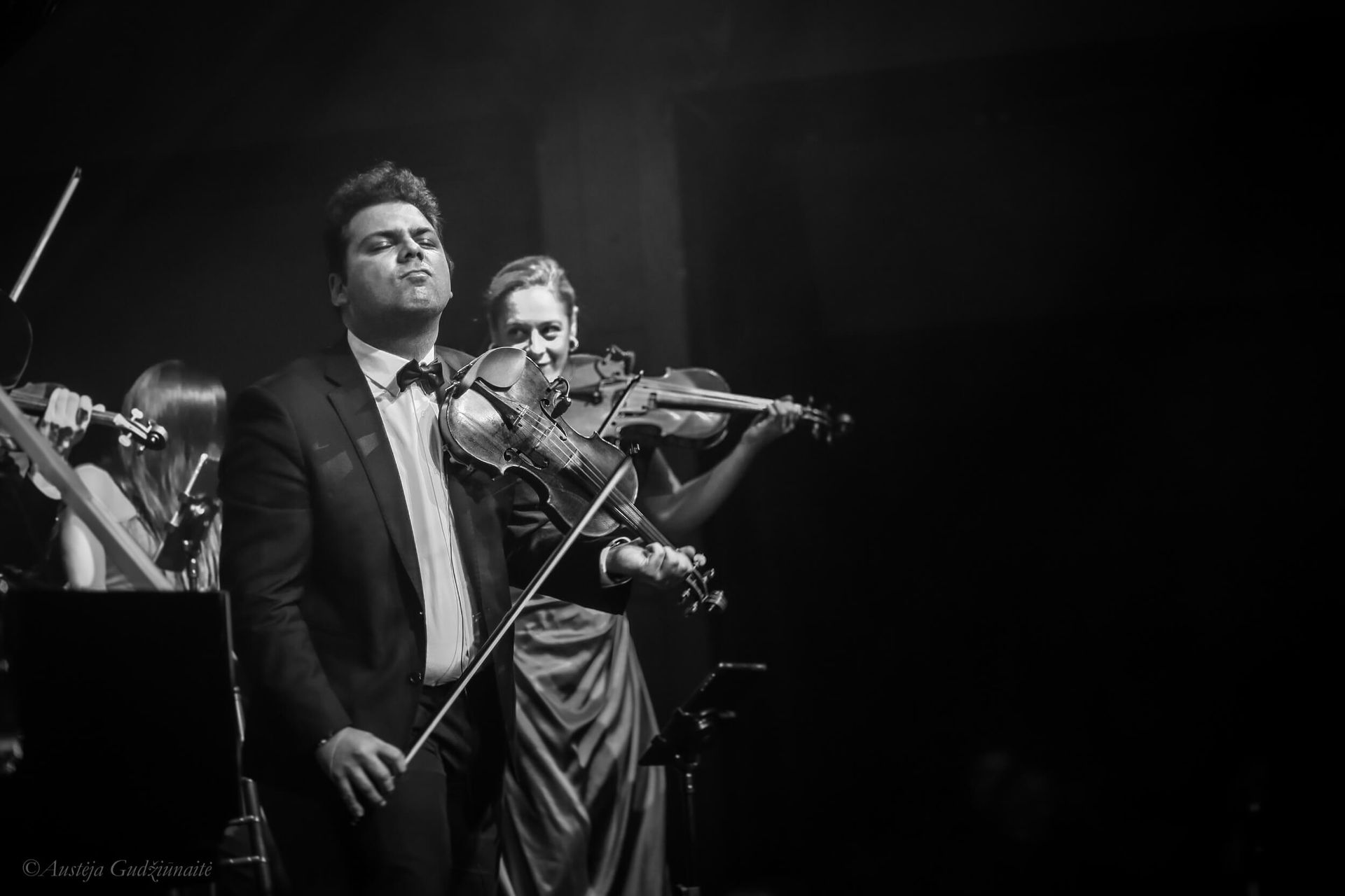 Musicians performing on stage; man in suit plays violin, another musician beside him. Black and white.