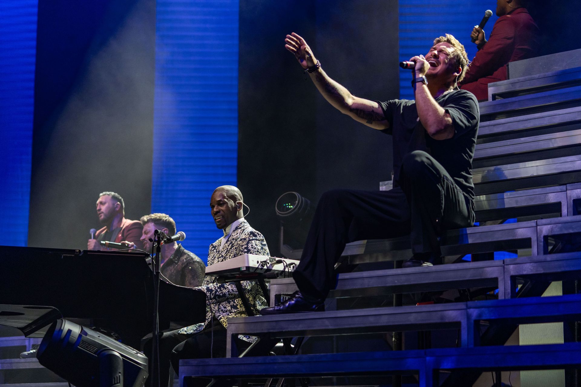 Singer on tiered stage, arms raised, performing with band, lit by blue and white lights.