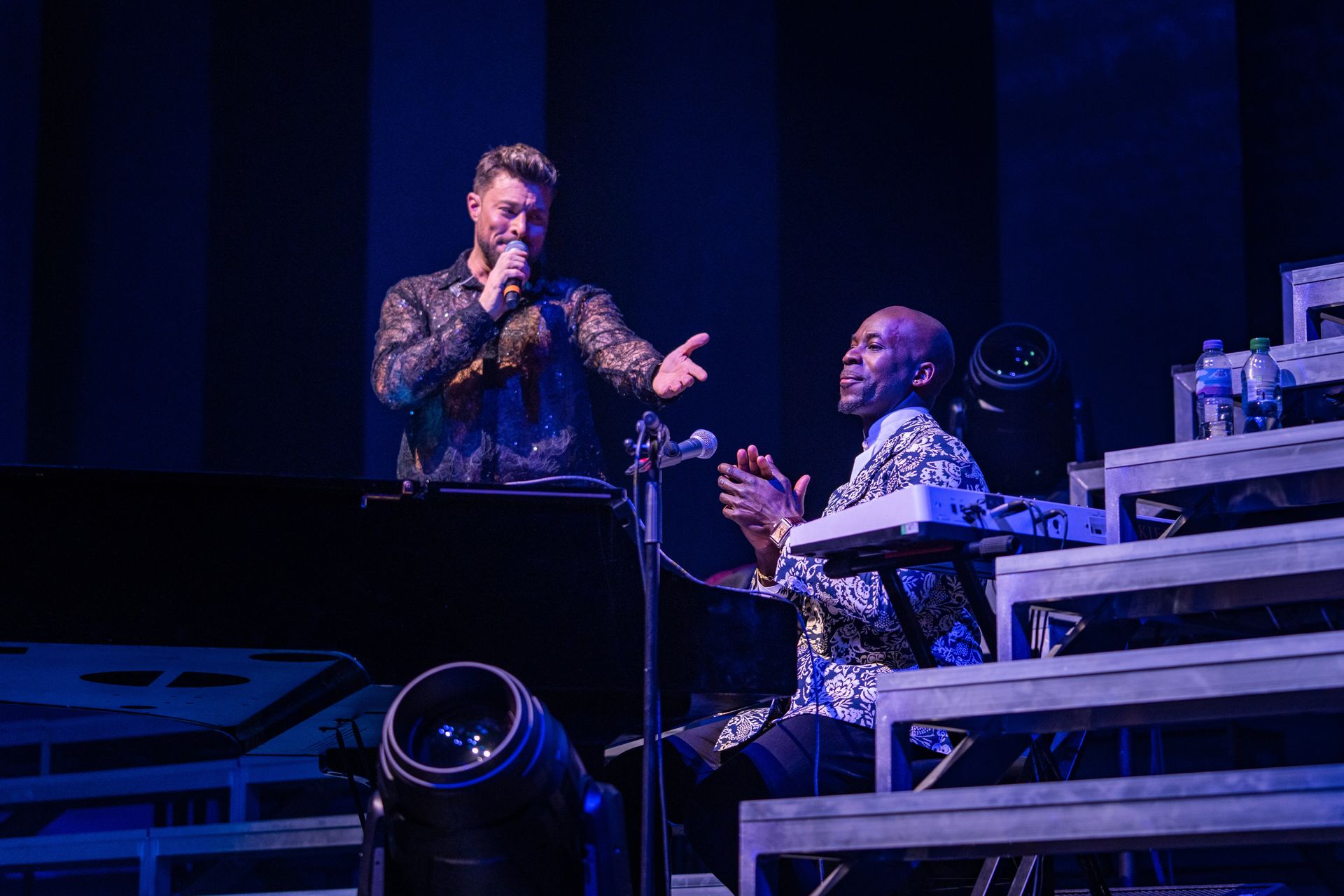 Two performers on stage: One singing, gesturing. The other playing a keyboard, clapping. Blue stage lighting.