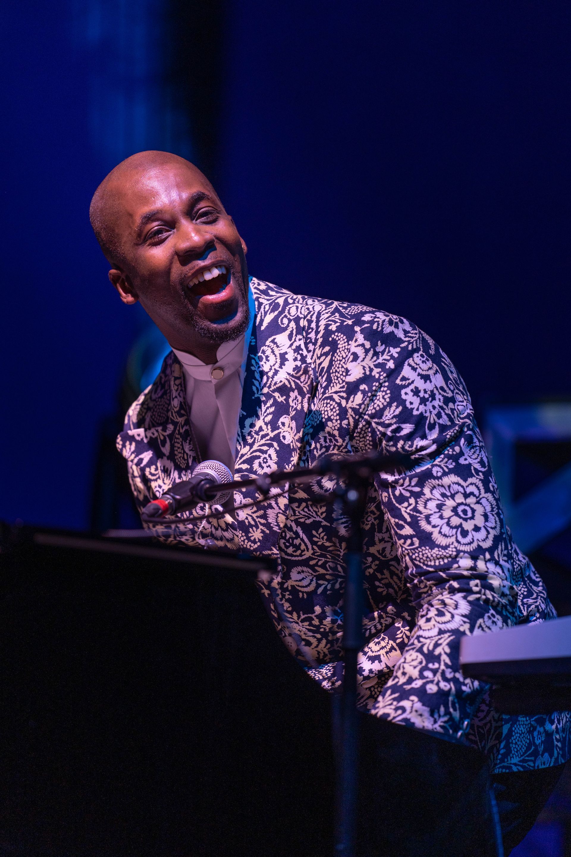 Man in white and blue patterned blazer leans over a keyboard, smiling, lit by blue stage light.