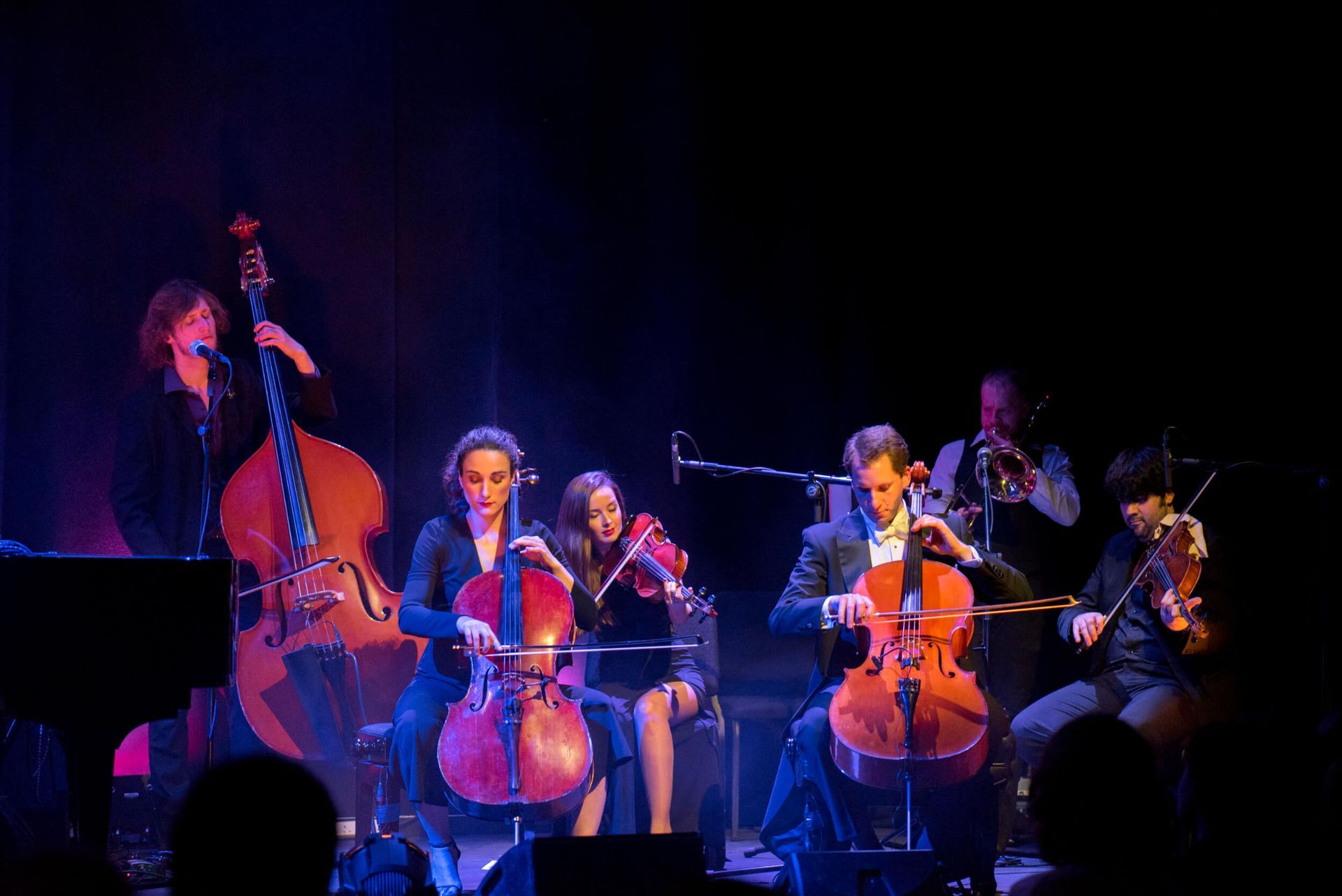 Musical ensemble on stage, featuring cello, violin, double bass, and trumpet, under stage lighting.