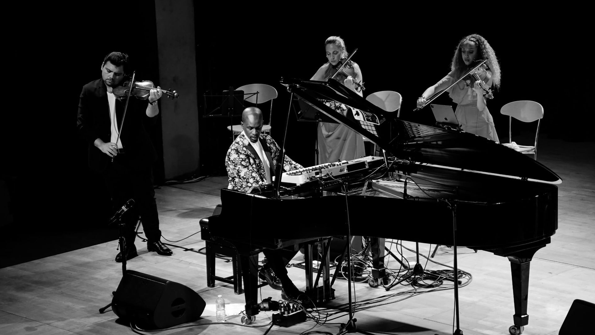 A jazz quartet performing on stage: piano, two violins, and a person playing a piano. Black and white.