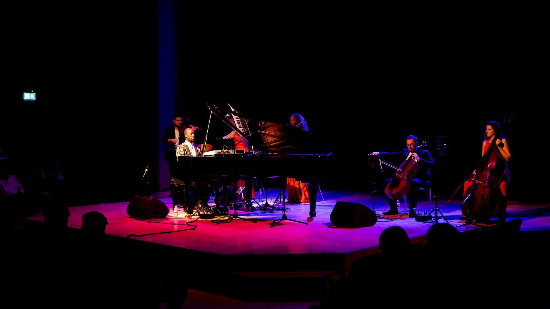 Musicians performing on a stage lit with red and blue. A grand piano is central, with cellos and a vocalist.