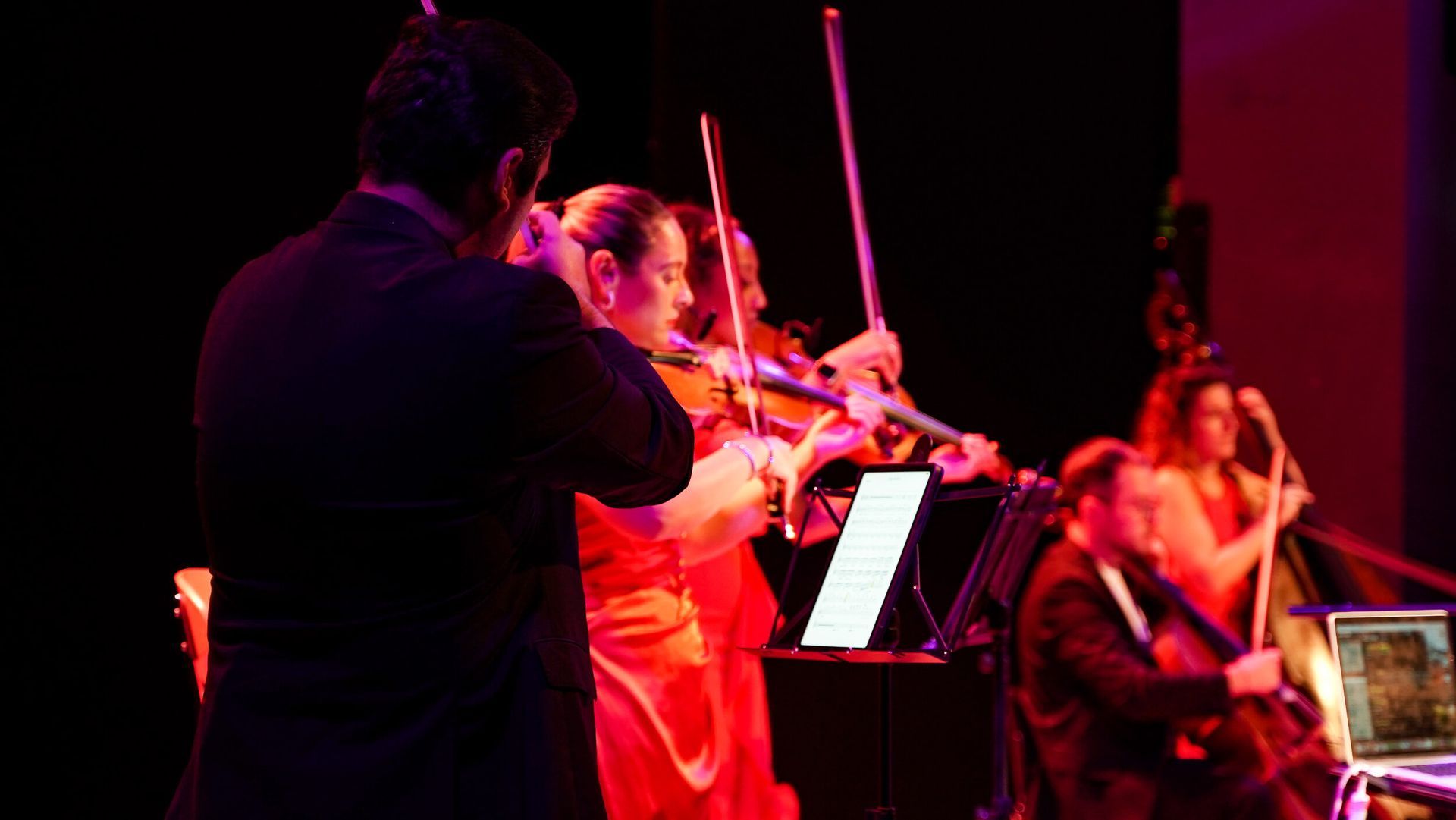 String quartet performing on stage, lit with red and purple lights. Musicians playing instruments, sheet music visible.