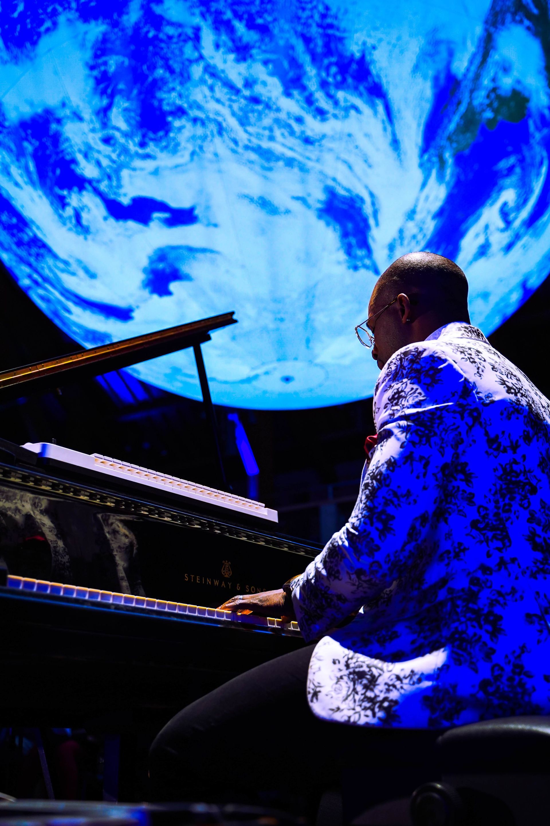 Pianist in a blue patterned jacket plays a grand piano, against a backdrop of a blurred Earth.