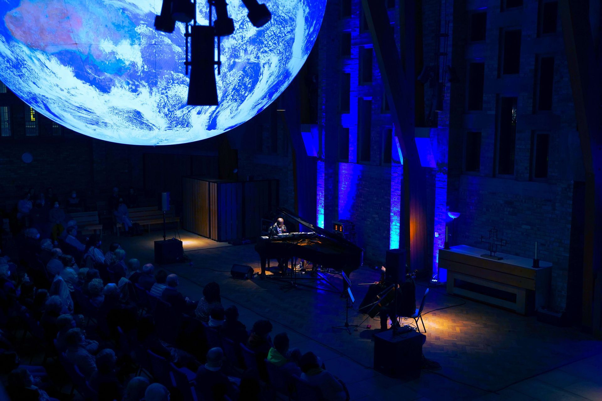 Concert with pianist and cellist on stage, Earth projected above. Audience in seats.