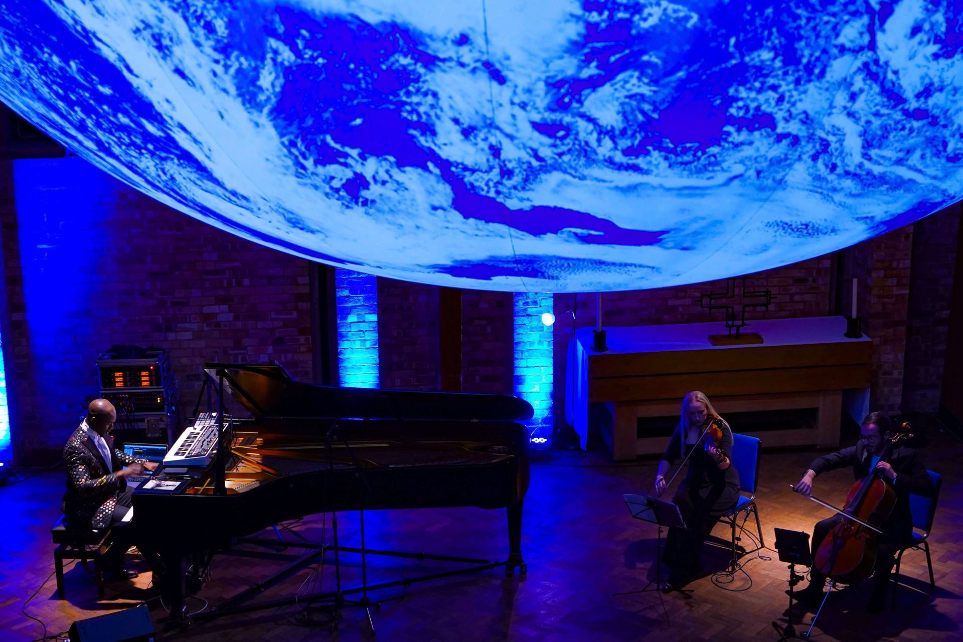 Musicians performing under a blue, Earth-like projection: piano, cello, and double bass.