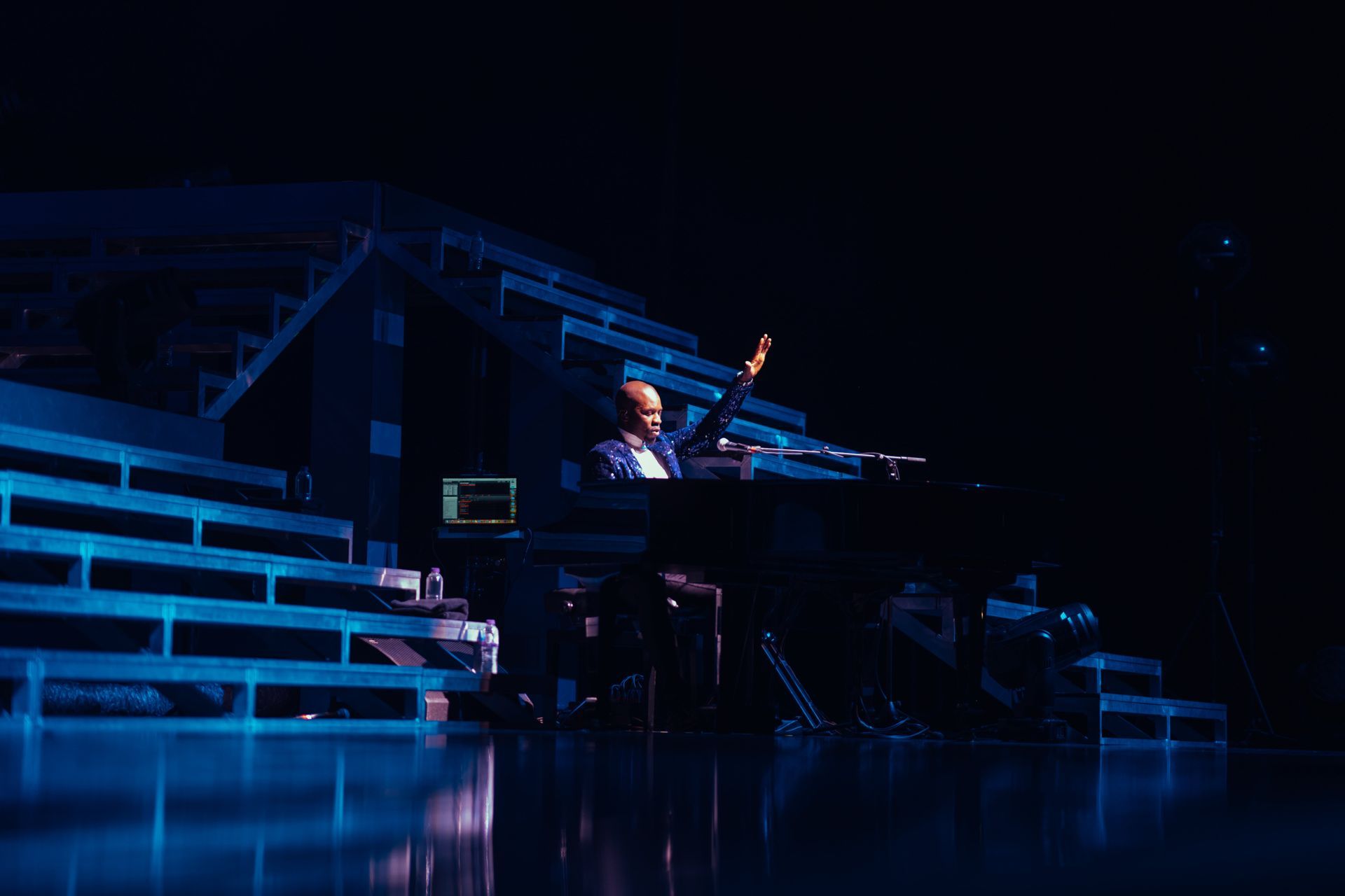 Man playing piano on stage, raising hand. Blue stage lighting.
