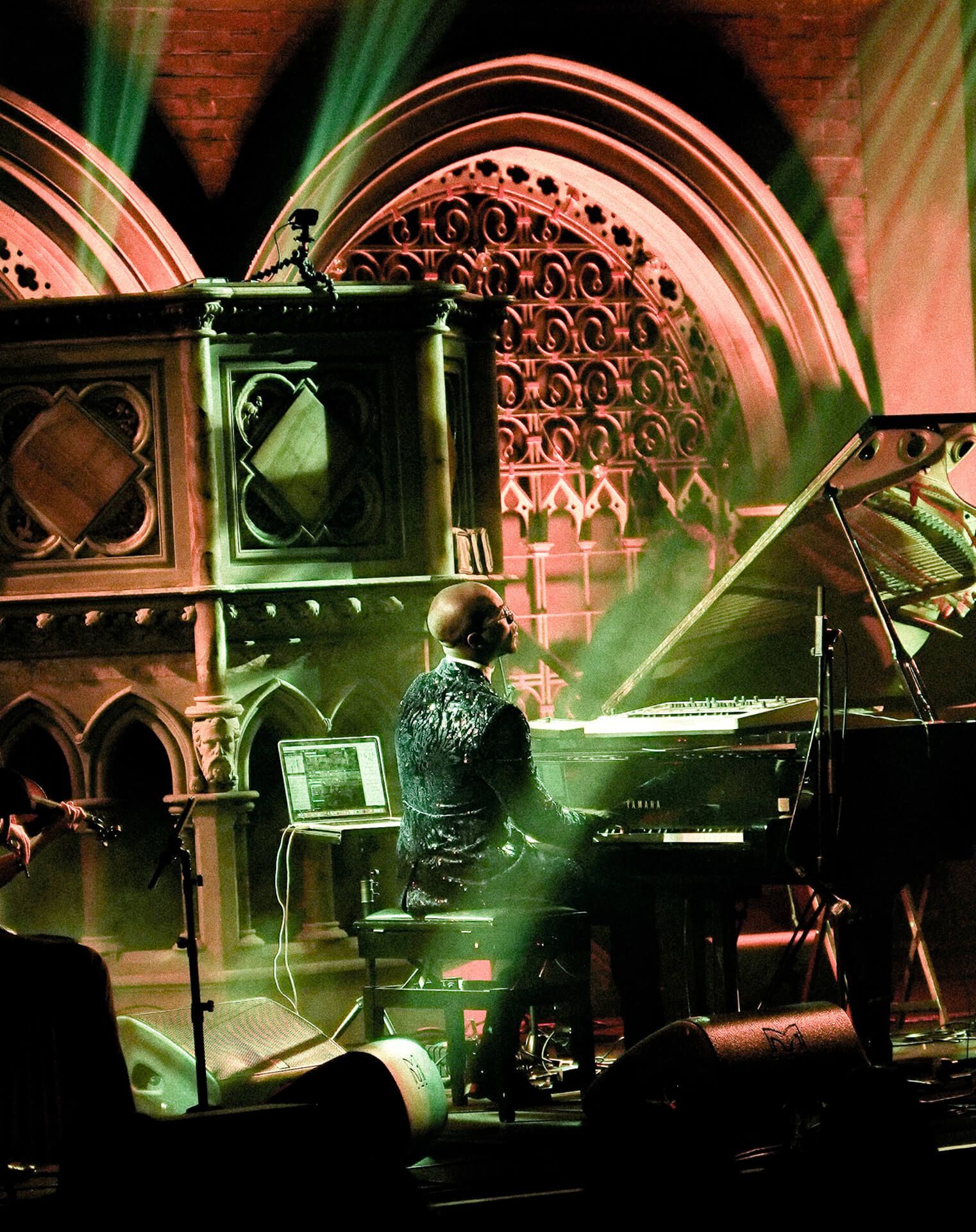 Person playing piano on a stage with a church-like backdrop, illuminated by green and red lights.