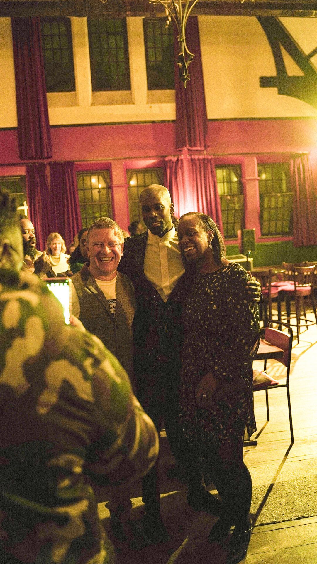 Three people smiling and posing for a photo in an ornate room with red curtains and windows.