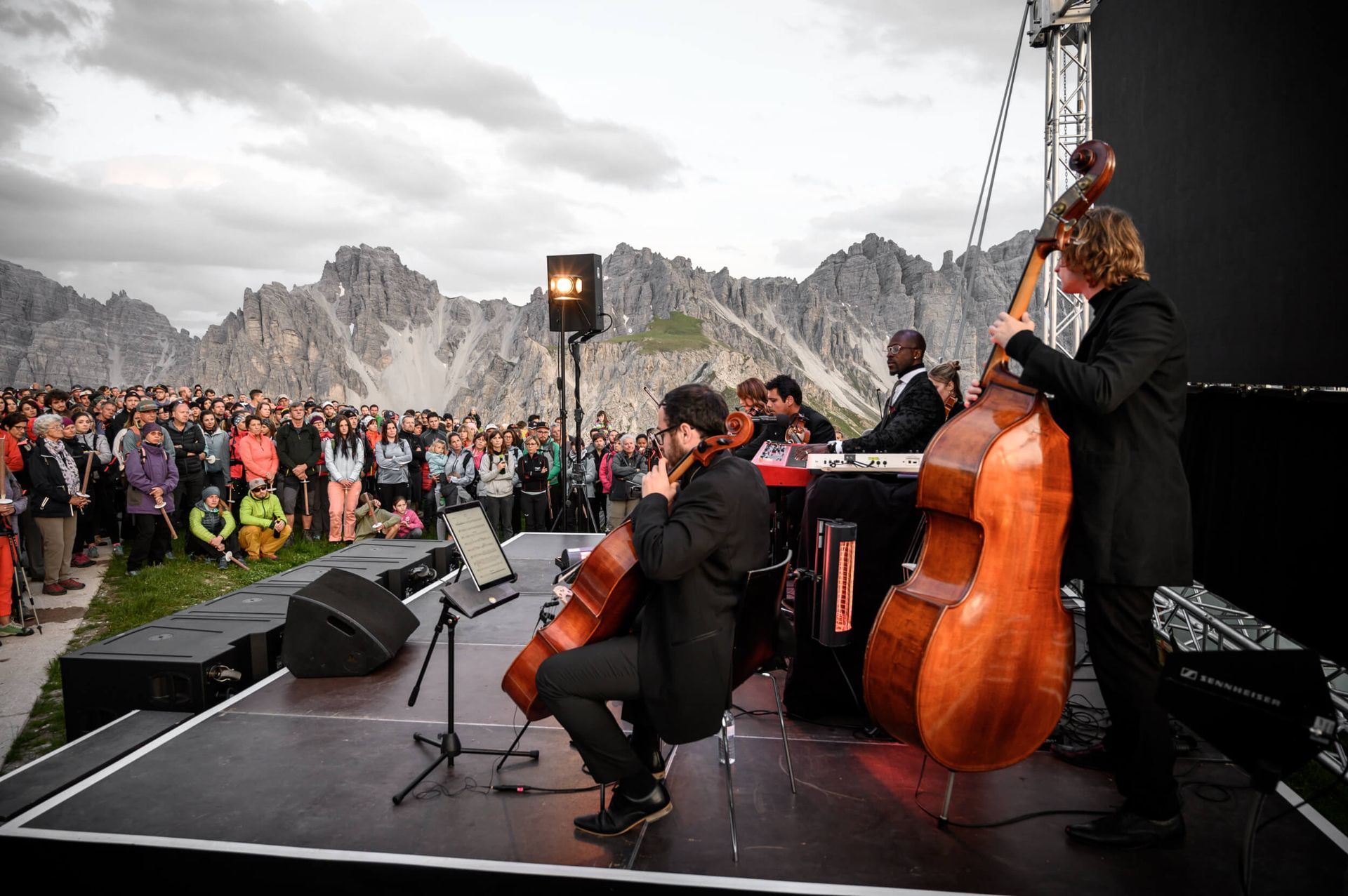 A string quartet performs on an outdoor stage with mountains in the background. A crowd watches the performance.