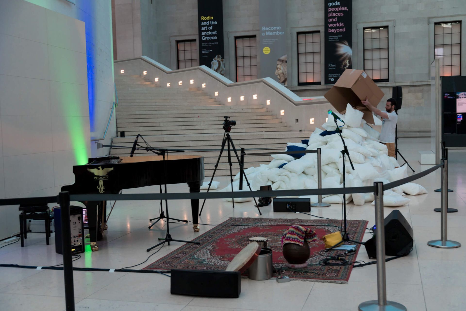 A person empties a box onto a pile of white pillows in a museum. A piano, microphone, and rug are in the foreground.