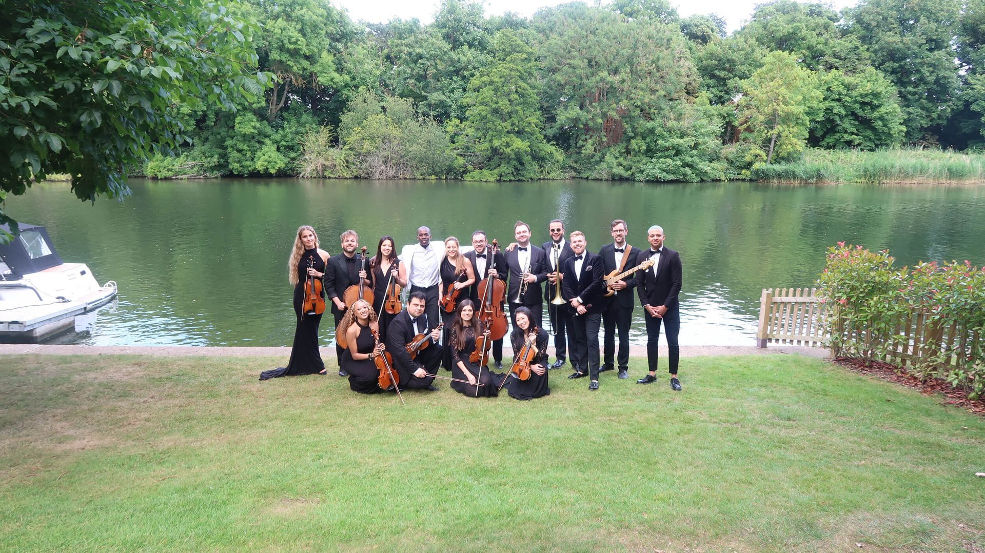Orchestra posing on a grassy bank beside a river. Musicians in formal attire; trees and boat in background.