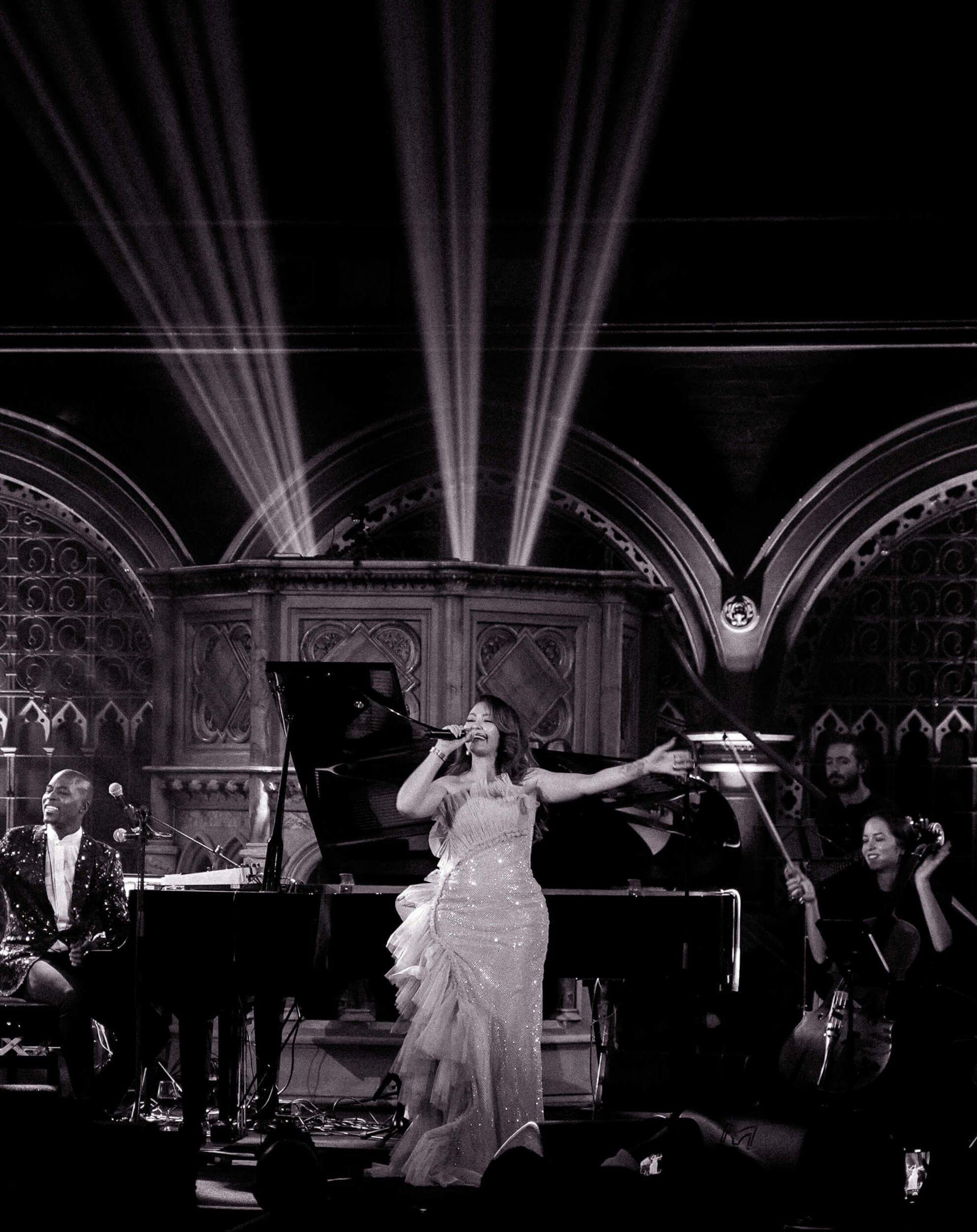 Singer in sparkling gown performs on stage with piano and string section.