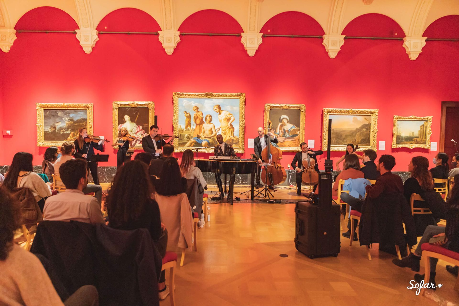 String quartet performs in a museum hall with audience. Red walls, paintings, and stage setup.