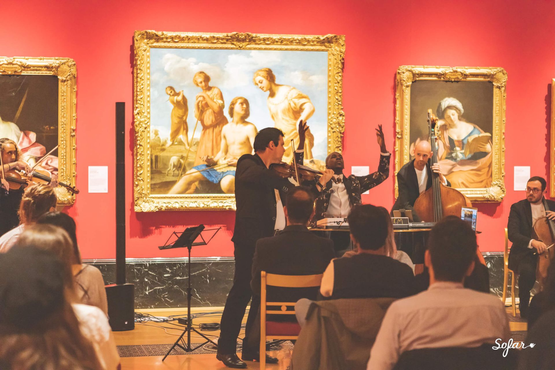 Musicians performing in a gallery with paintings on red walls. Audience seated, listening.