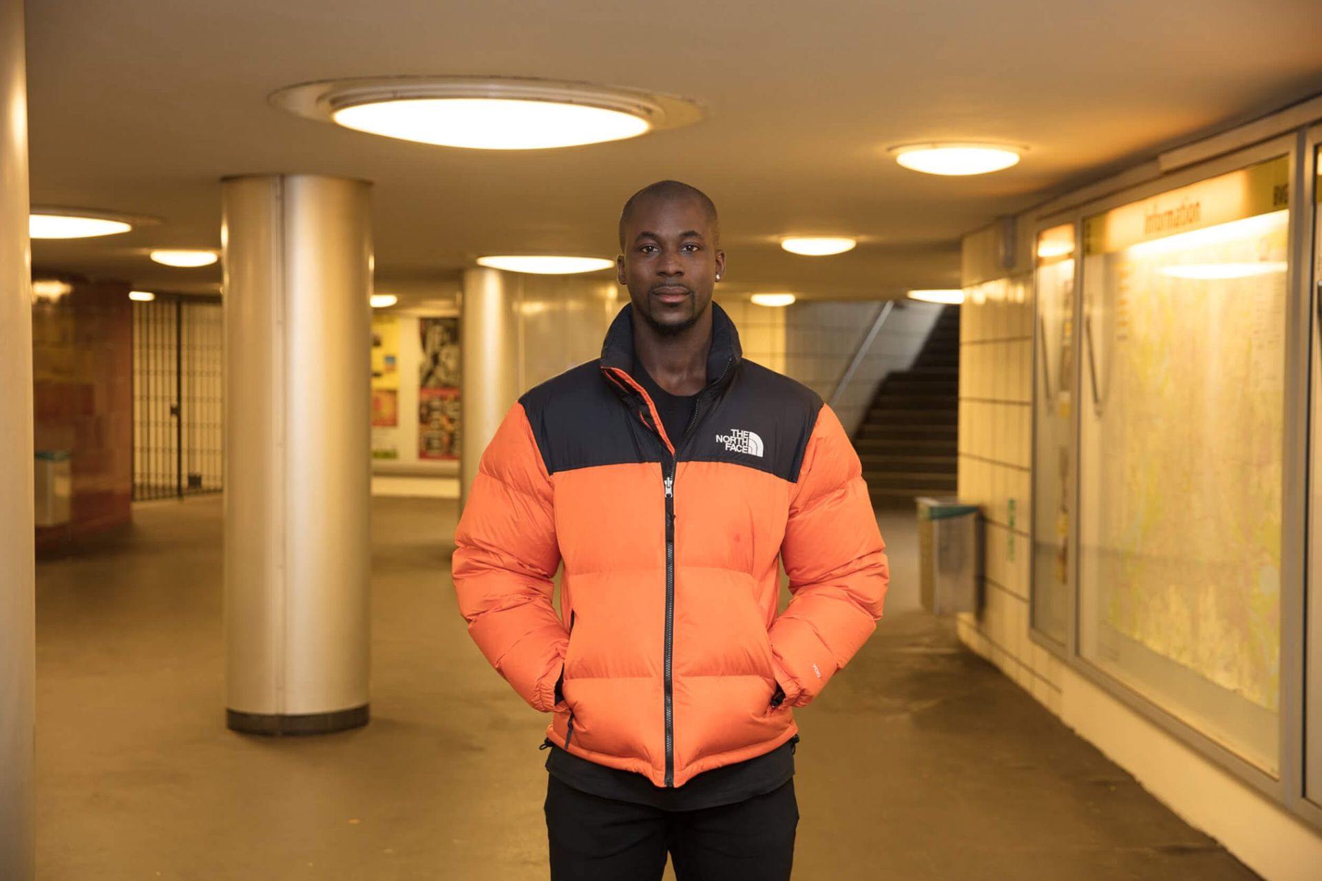 Man wearing an orange and black puffer jacket stands in a subway station, hands in pockets.