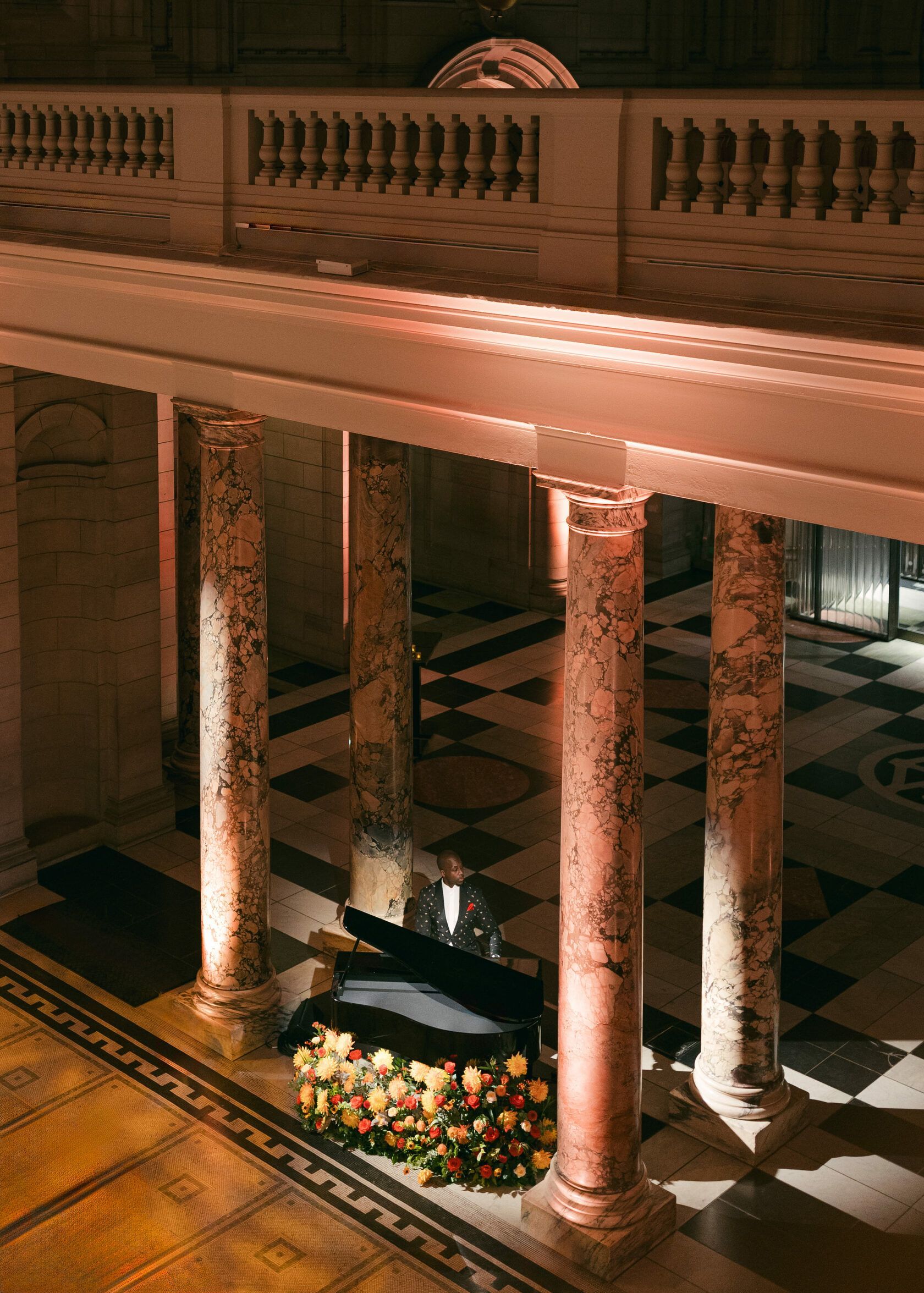 Grand piano beneath marble columns, lit by warm lighting, set in a formal hall with patterned floor and balcony above.