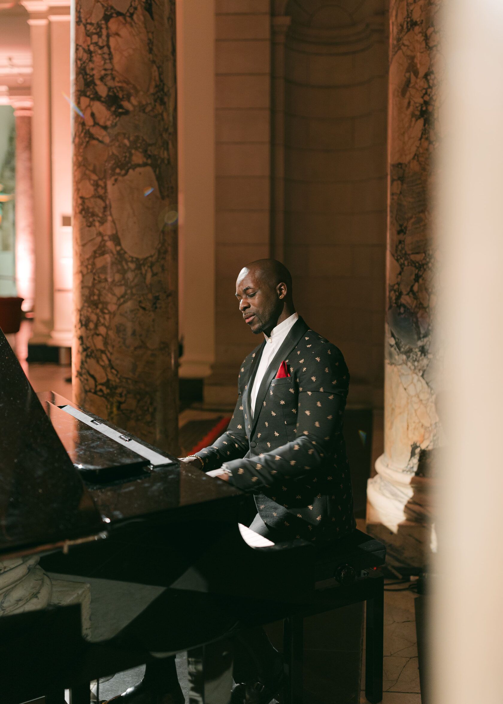 Man in dotted blazer playing a grand piano indoors. Elegant setting, columns, sheet music.