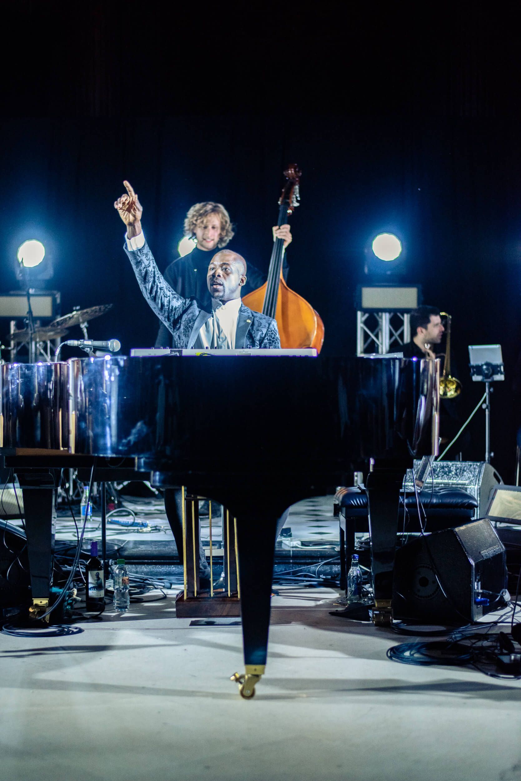 A pianist in a sparkly suit raises an arm. Bandmates stand behind him with a bass and horn on stage.