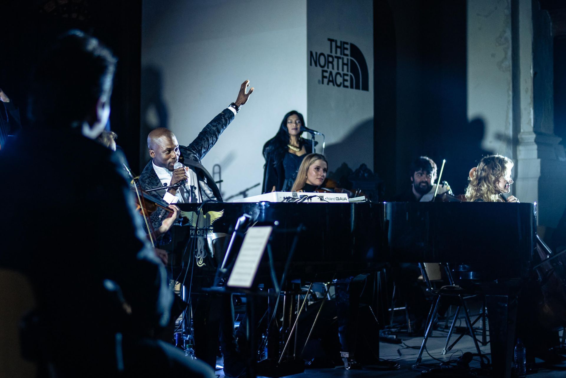 A band performing live with a piano and string section; The North Face logo in the background.