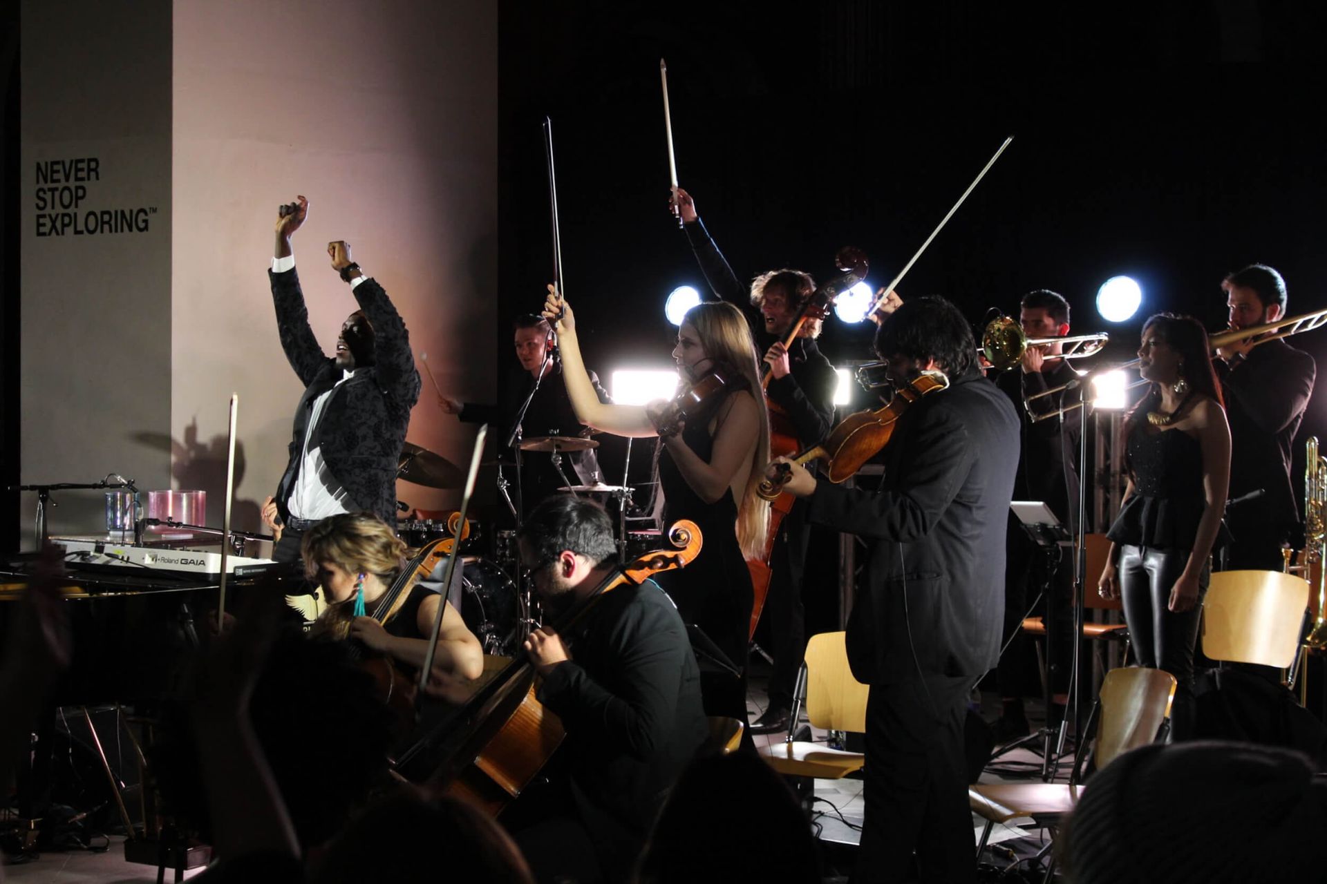 Orchestra performing on stage; conductor raises arms, musicians play. Dim lighting, spotlights.