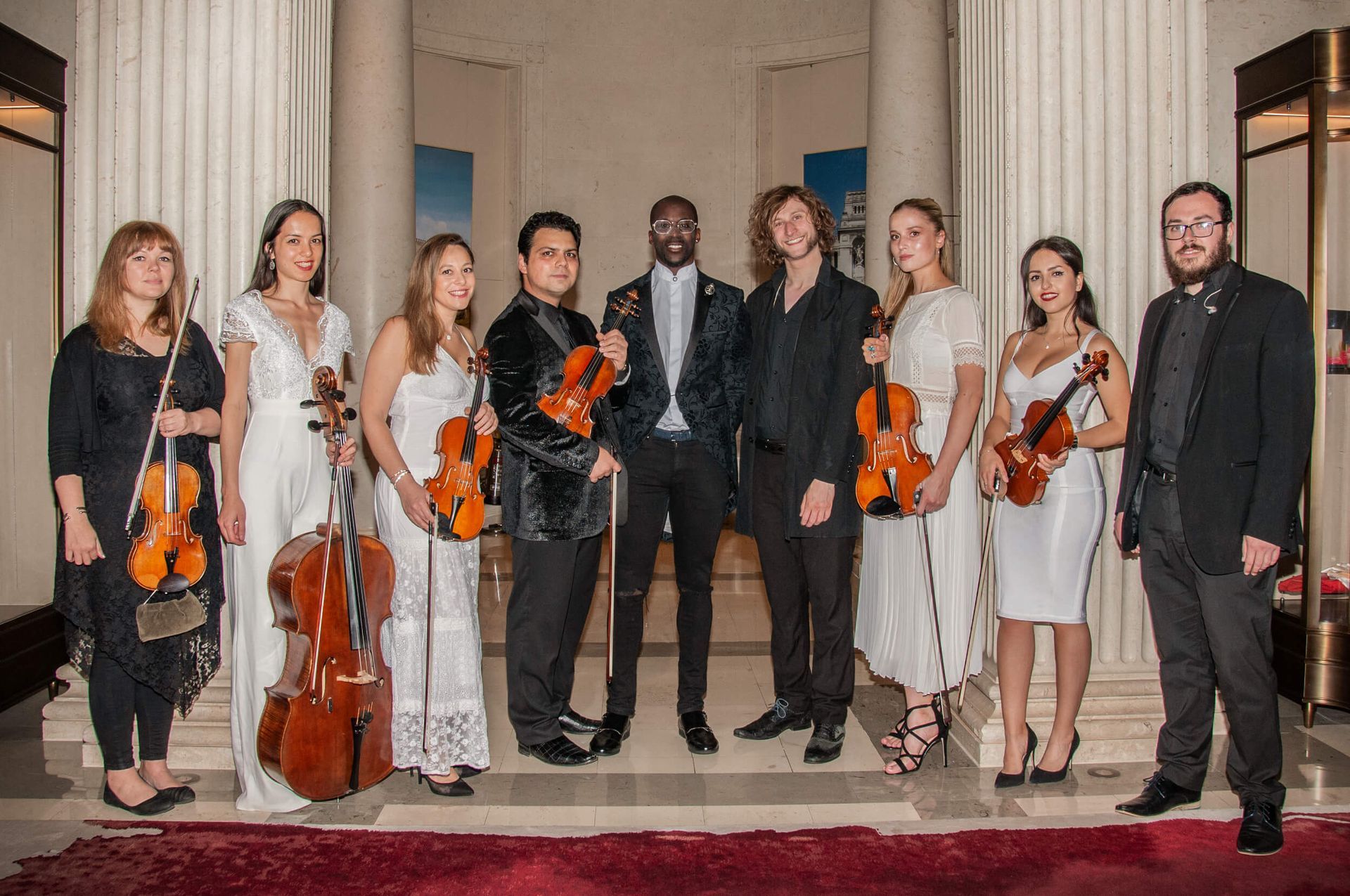 A string ensemble of nine musicians posing indoors near white pillars and a red rug.