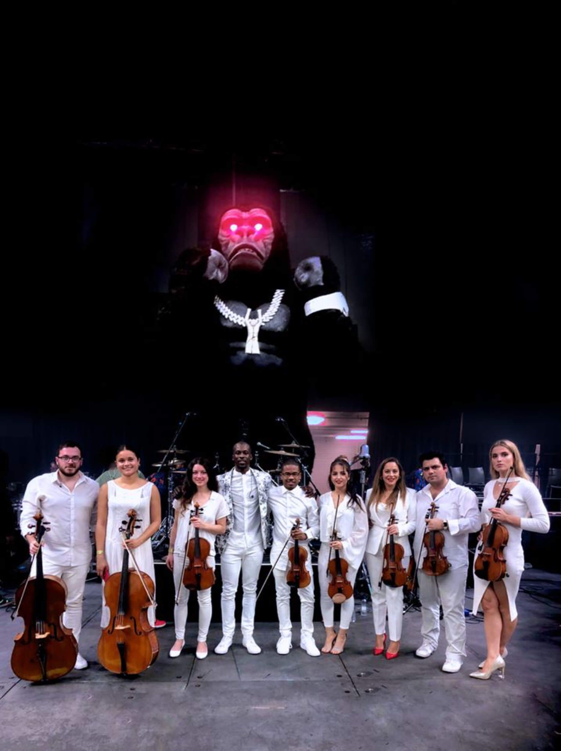 Orchestra in white outfits, holding instruments, performing on stage with a dark background and a projection.