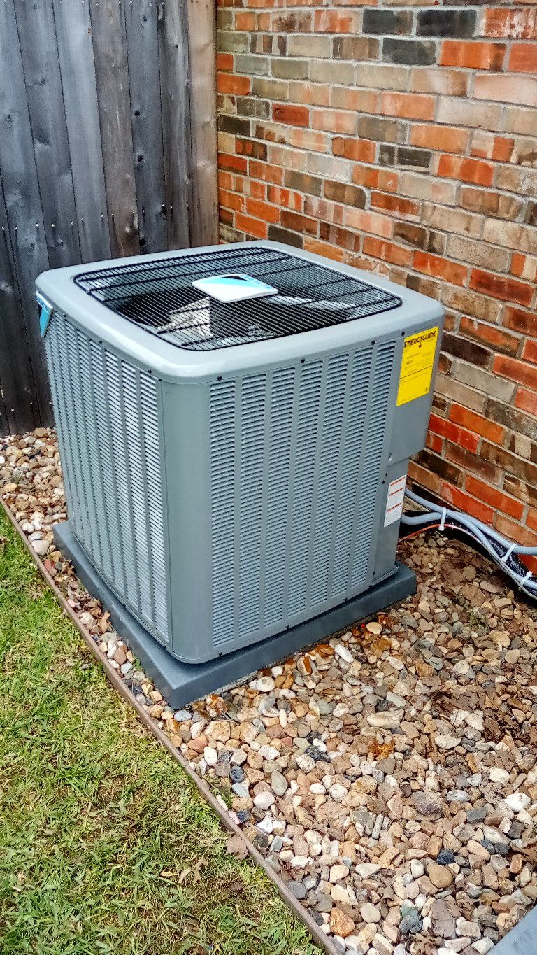 Air conditioning unit on a concrete base, next to a brick wall and wooden fence.
