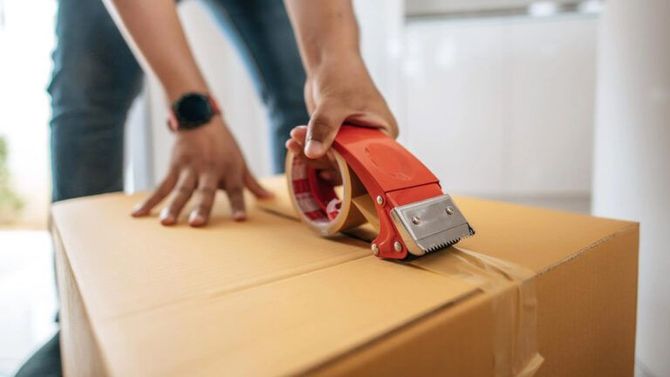 A person uses a red tape dispenser to seal a cardboard moving box.