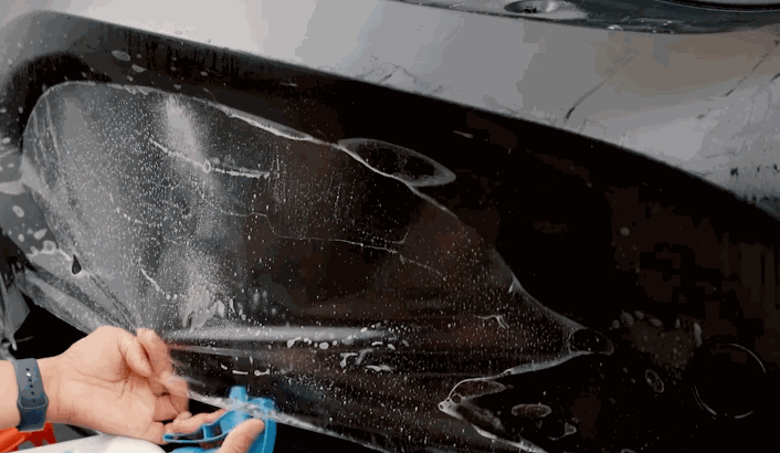 Hands trimming and adjusting paint protection film on a black rear bumper, with visible soapy water underneath the film.