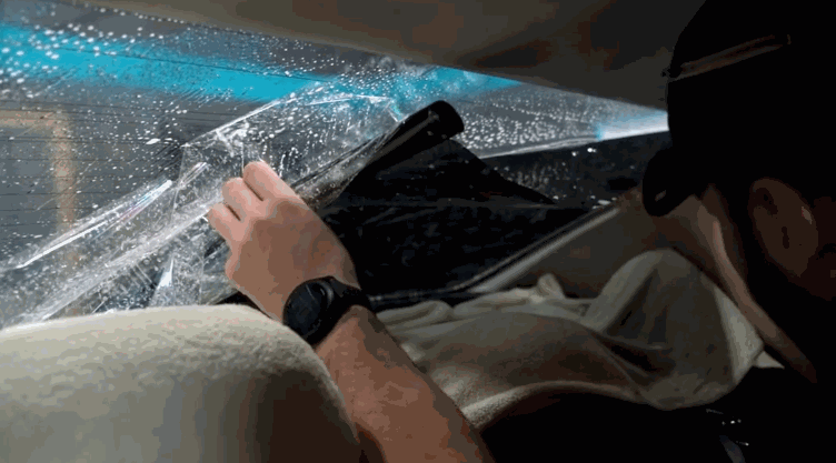 An installer carefully positions window tint film on the inside of a car’s rear window using a squeegee and heat for alignment.