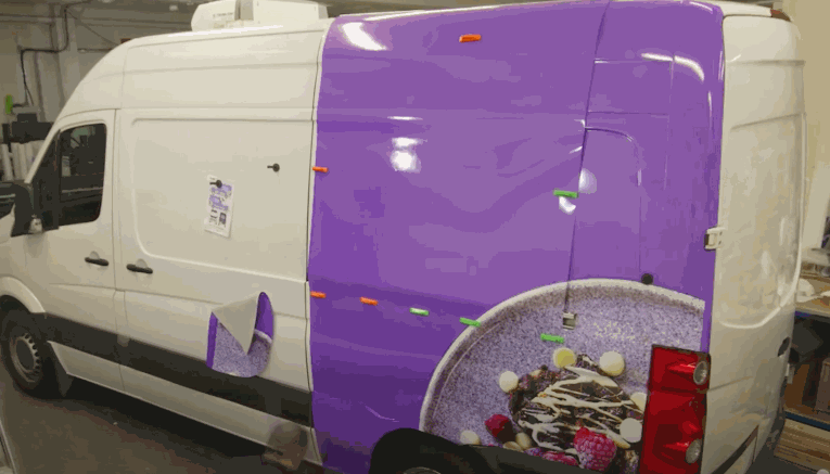 A white van is partially wrapped with a vibrant purple vinyl graphic featuring a food image, held in place with colorful clips as the wrap is carefully aligned and applied.