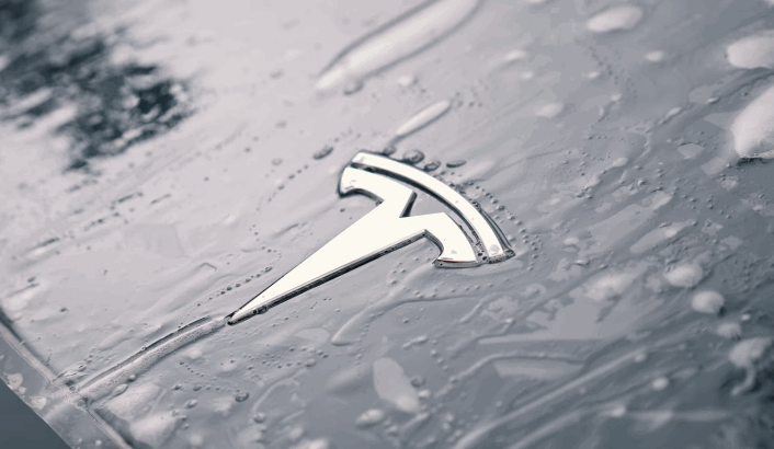 Tesla logo on a wet car hood with paint protection film partially applied, showing water droplets trapped beneath the surface.