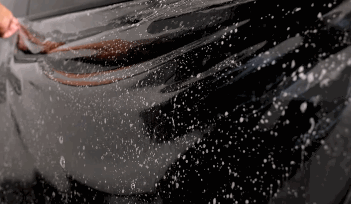 Close-up of a hand applying a wet paint protection film (PPF) sheet onto a glossy black car door with water bubbles underneath.